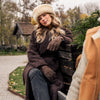 Model sitting on outdoor bench in park wearing brown sheepskin gloves and sheepskin hat
