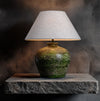 Table lamp with a textured green base and white shade on a stone surface against a dark background