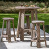 Outdoor Gommaire bar table with four stools made from teak wood, set on a concrete surface with a grassy background and trees in the distance.