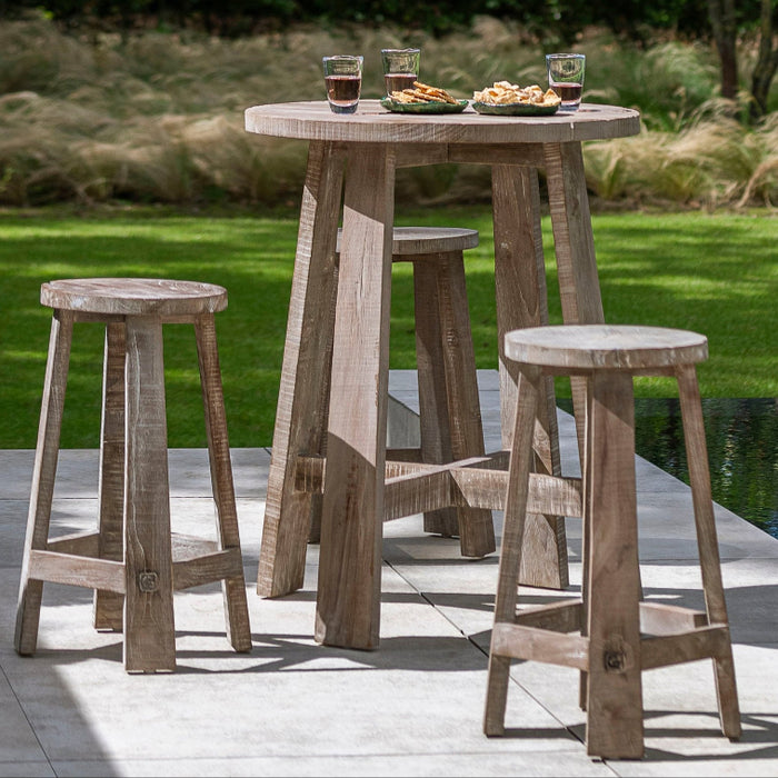 Outdoor Gommaire bar table with four stools made from teak wood, set on a concrete surface with a grassy background and trees in the distance.