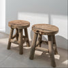 Two solid teak stools against a white interior wall on tiled floor