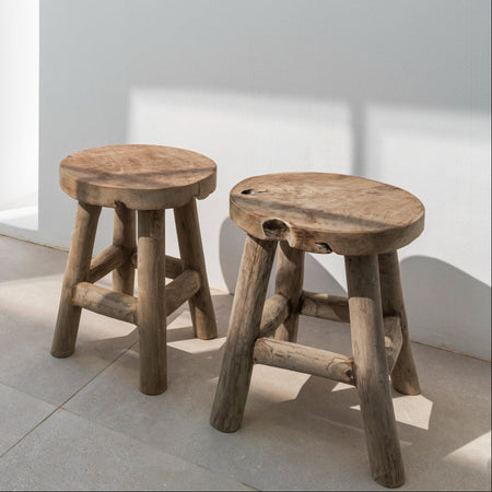Two solid teak stools against a white interior wall on tiled floor