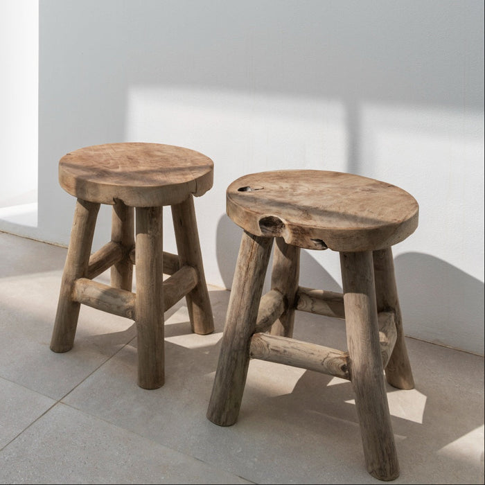 Two solid teak stools against a white interior wall on tiled floor