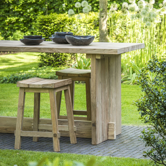A wooden bar table with a small stool beside it, placed on a garden pathway, with a bowl and glassware on the table.