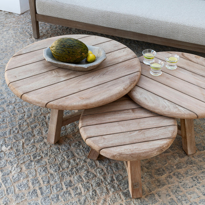 Trio of round teak coffee tables by Gommaire in a natural grey finish, placed on a tiled floor next to a beige sofa with a bowl of fruit and three small glasses.