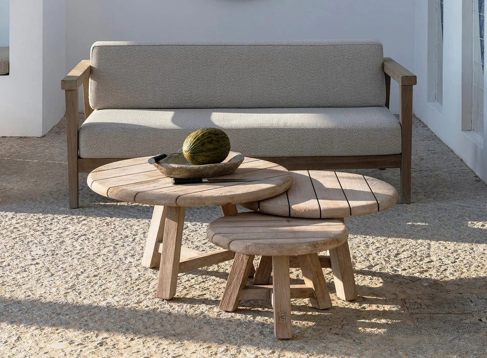 Three teak coffee tables with a natural grey patina in small, medium and large sizes on a stone terrace with sofa and whitewashed wall behind.