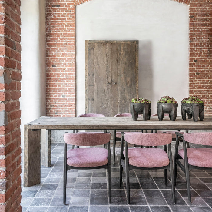 A large rectangular dining table made from reclaimed wood with an aged patina, positioned in a room with exposed brick walls and a wooden door.