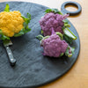 Circular black wooden chopping or serving board with knife and coloured brassicas on a wooden table