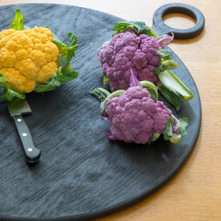 Circular black wooden chopping or serving board with knife and coloured brassicas on a wooden table