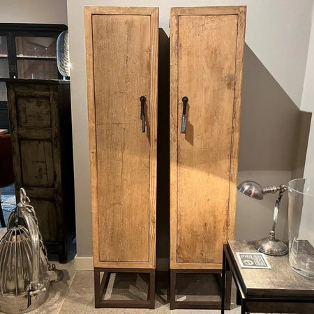 Two tall wooden cabinets in a room with a table and lamp.