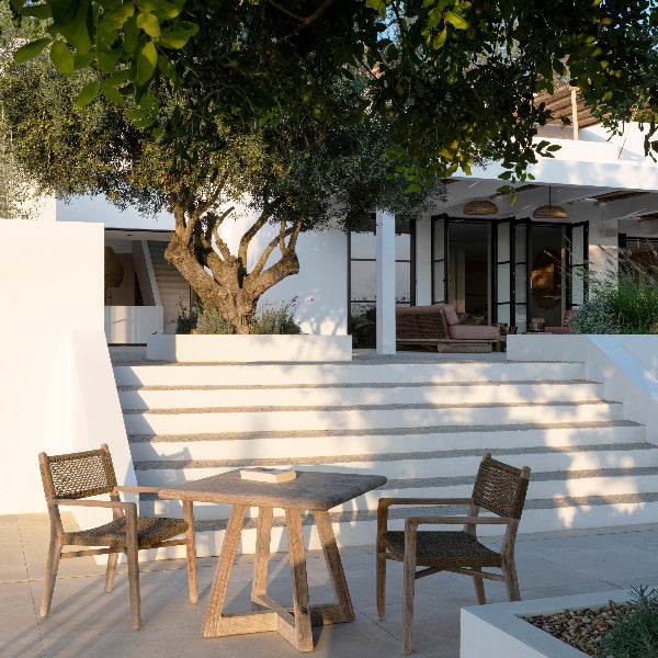 A bistro table made of teak wood with a natural grey patina, featuring a crossed leg configuration, displayed in an outdoor setting with a house in the background.