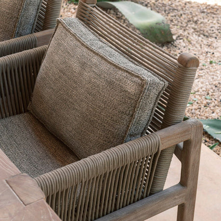 Close up of natural teak dining armchair with rope weave back and arms next to a table on outdoor terrace