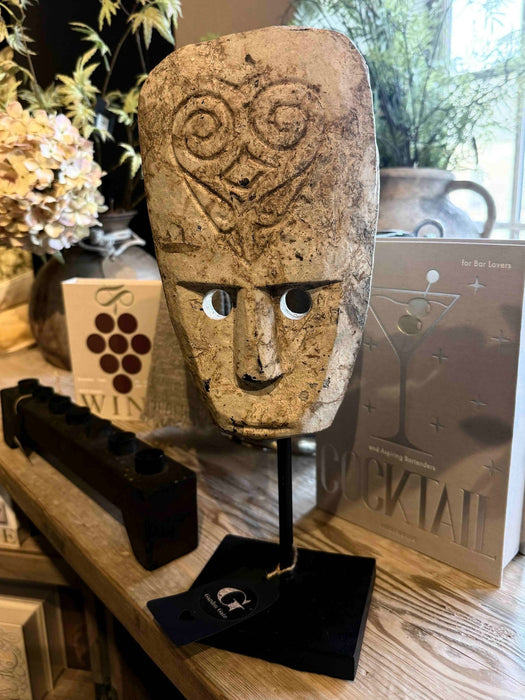 Beige tribal carved mask on black stand sitting on wooden shelf