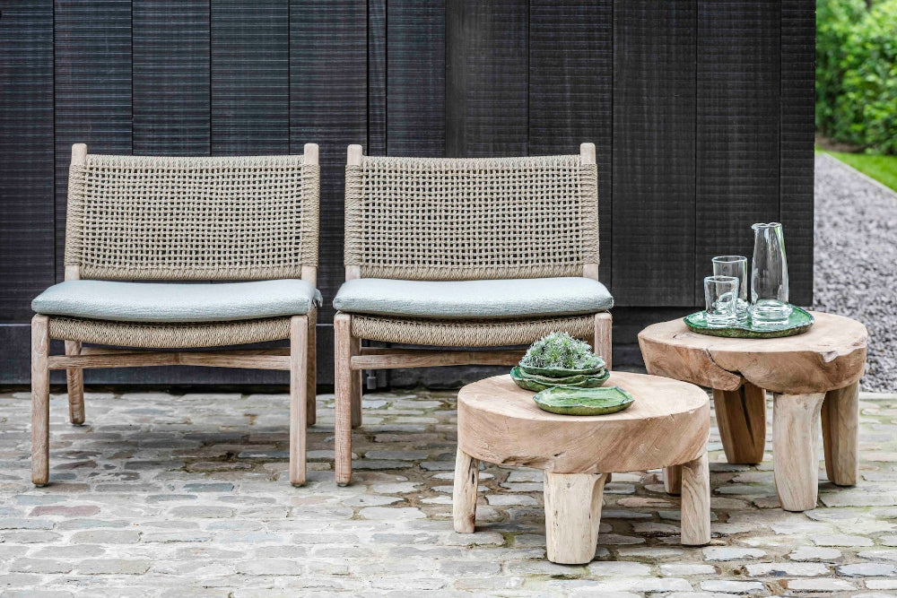Two reclaimed teak and wicker easy chairs by Gommaire with cushion in front of dark wood fence on brick paved area with two low teak Gommaire Samba tables