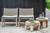 Two reclaimed teak and wicker easy chairs by Gommaire with cushion in front of dark wood fence on brick paved area with two low teak Gommaire Samba tables