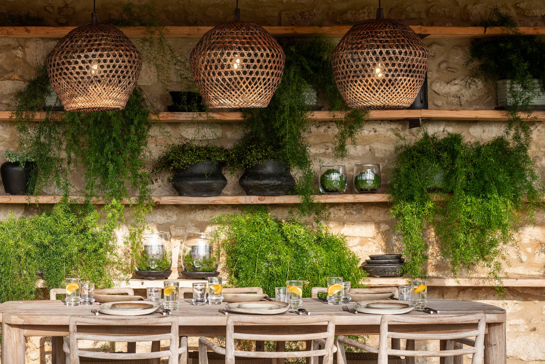 Dining area with rustic shelves, hanging lights, and greenery against a stone wall.
