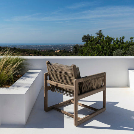 A teak chair with natural grey rope seat and backrest, placed on a white surface with a landscape in the background.