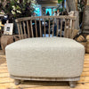 A beige upholstered lounge seat with a wooden frame, featuring a cushioned seat and curved backrest, placed on a wooden floor with plants in the background.