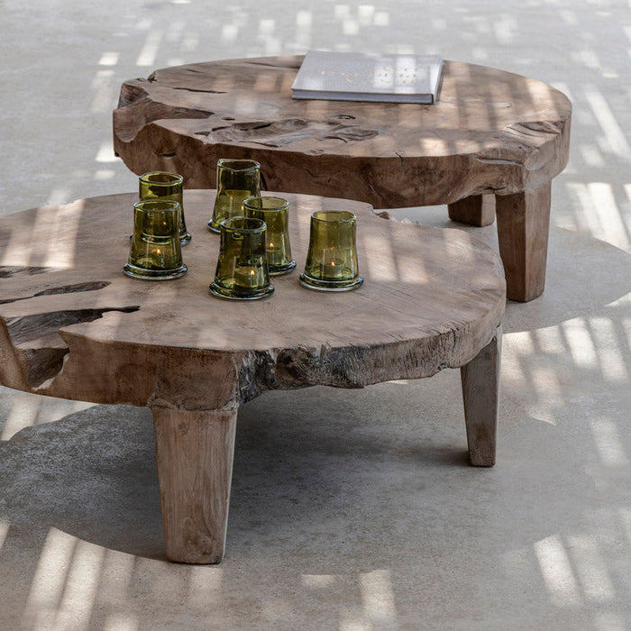 A low, round, natural grey finish wooden coffee table placed on a concrete floor, with bottles on top of it and shadow patterns on the surface.