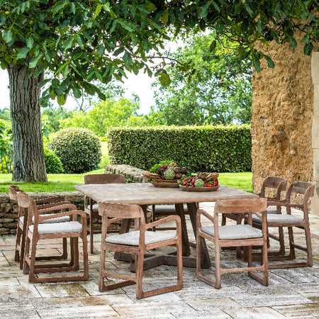 A set of teak armchairs with beige cushions arranged on a patio next to a dining table, with a garden and a house in the background 
