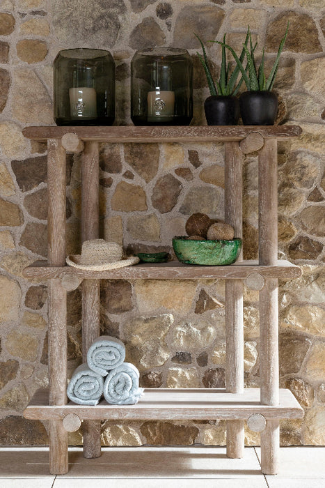 A teak shelving rack with three tiers, mounted on a stone wall. It is filled with various decorative items including potted plants and a beige blanket.