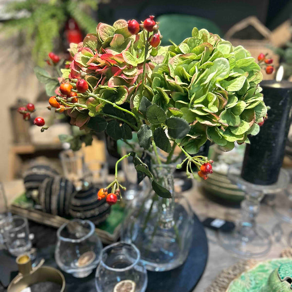 Decorative floral arrangement with green leaves and red berries in a clear vase on a table