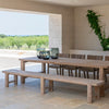 A long teak dining bench by Gommaire with a natural grey patina, placed on a tiled floor, with a table in the background, set in an outdoor or covered patio area with a scenic view.