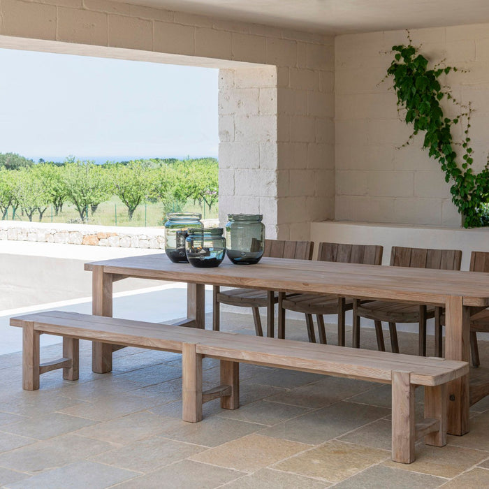 A long teak dining bench by Gommaire with a natural grey patina, placed on a tiled floor, with a table in the background, set in an outdoor or covered patio area with a scenic view.