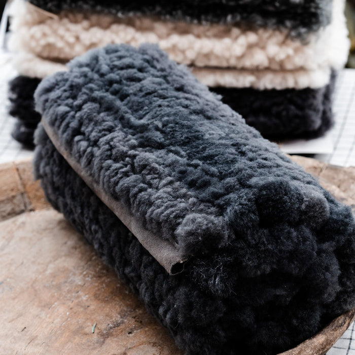 Three oblong patchwork sheepskin bench cushions in black, gray, and white, displayed on a wooden surface with a checkered pattern in the background.