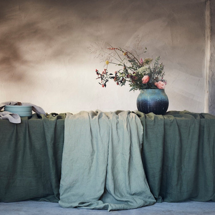 Pale green linen tablecloth with darker green linen tablecloths draped over a table with floral arrangement, pile of bowls and a rustic wall background.