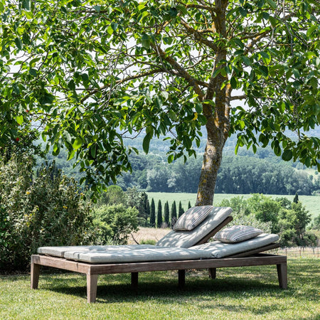 Outdoor teak daybed with cushions under a tree in a garden setting