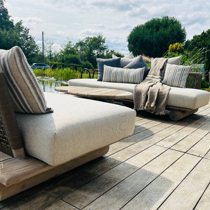 Pair of luxury teak outdoor sofas with deep cream base cushions, decorative striped cushions and teak outdoor coffee tables on decking next to pond