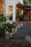 A series of outdoor LED pillar candles lined up on a pathway, with a house and plants in the background.