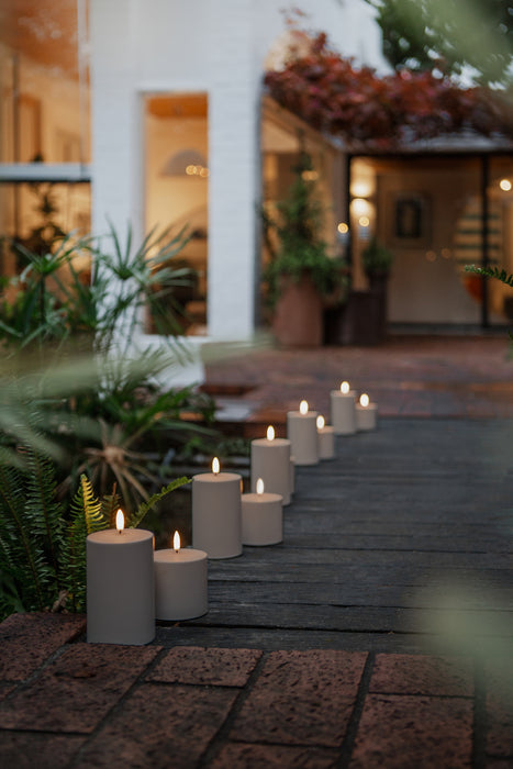 A series of outdoor LED pillar candles lined up on a pathway, with a house and plants in the background.