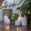 Three white LED pillar candles placed on a brick pathway with plants in the background.
