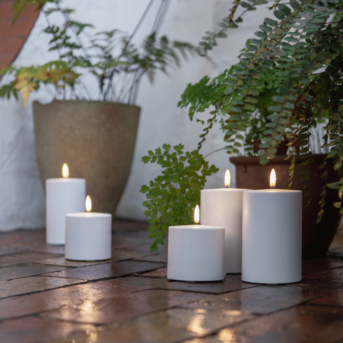 Three white LED pillar candles placed on a brick pathway with plants in the background.