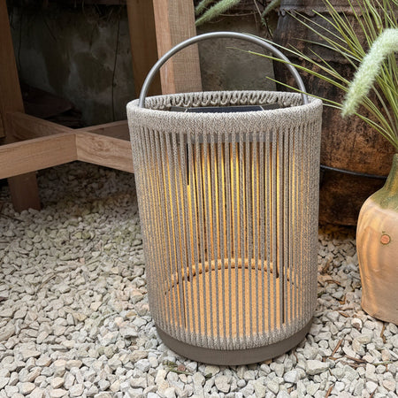 A cylindrical woven rope lantern in a light beige/taupe color with a dark metal top handle, placed on a bed of white gravel next to a potted plant.