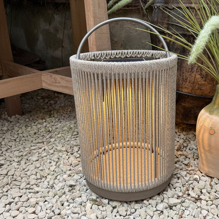 A cylindrical woven rope lantern in a light beige/taupe color with a dark metal top handle, placed on a bed of white gravel next to a potted plant.