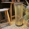 A cylindrical solar lantern in natural rope in a warm taupe-beige color, situated on a bed of white gravel next to a wooden bench and green pampas grass.