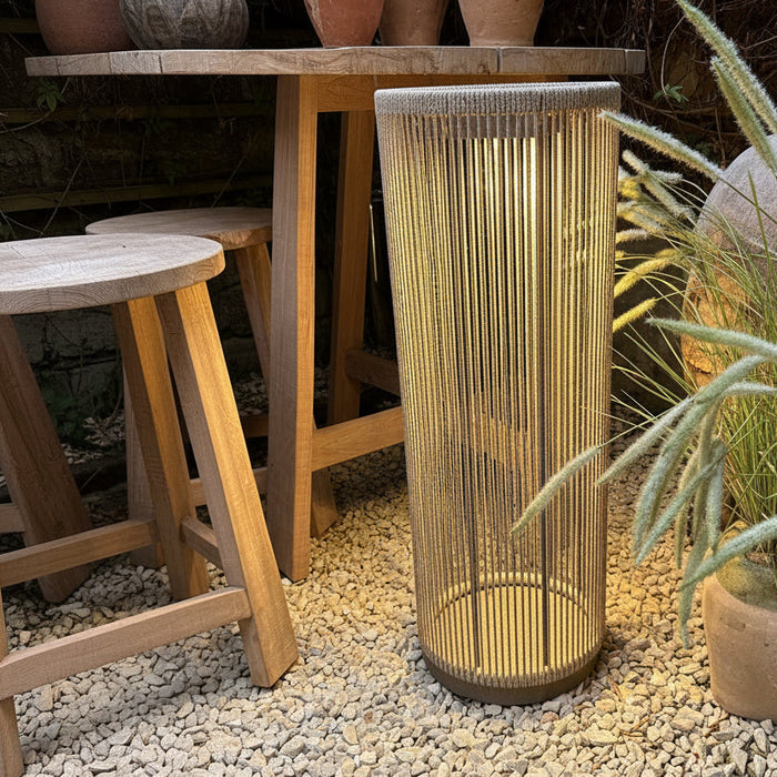 A cylindrical solar lantern in natural rope in a warm taupe-beige color, situated on a bed of white gravel next to a wooden bench and green pampas grass.