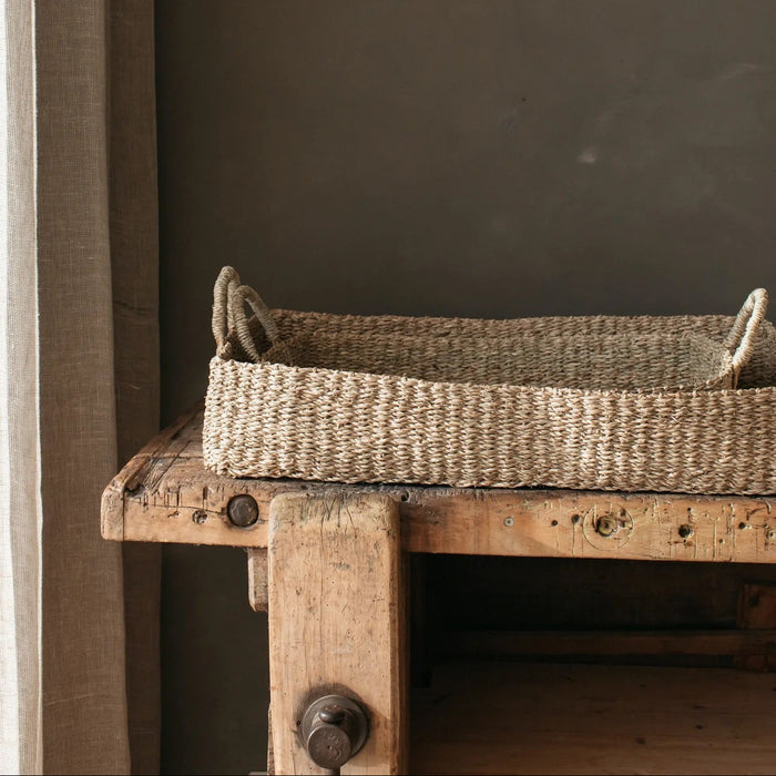 A small shallow seagrass basket with plaited handles, resting on a wooden surface against a textured wall.