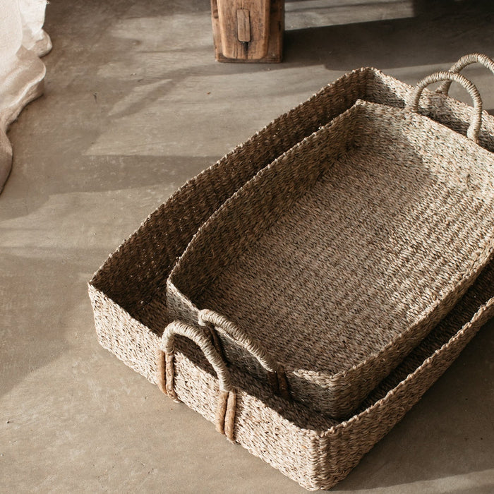 Two matching shallow seagrass baskets, one larger, one smaller inside pictured on a stone floor