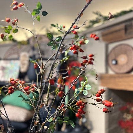 Artificial twig with small russet leaves pictured with faux red hip spray in floral arrangement with home decor items behind