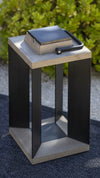 A modern square outdoor solar lantern with black metal frame and teak wood top and base with square solar light on top, set on a dark textured rug with green foliage in the background.