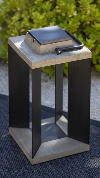 A modern square outdoor solar lantern with black metal frame and teak wood top and base with square solar light on top, set on a dark textured rug with green foliage in the background.