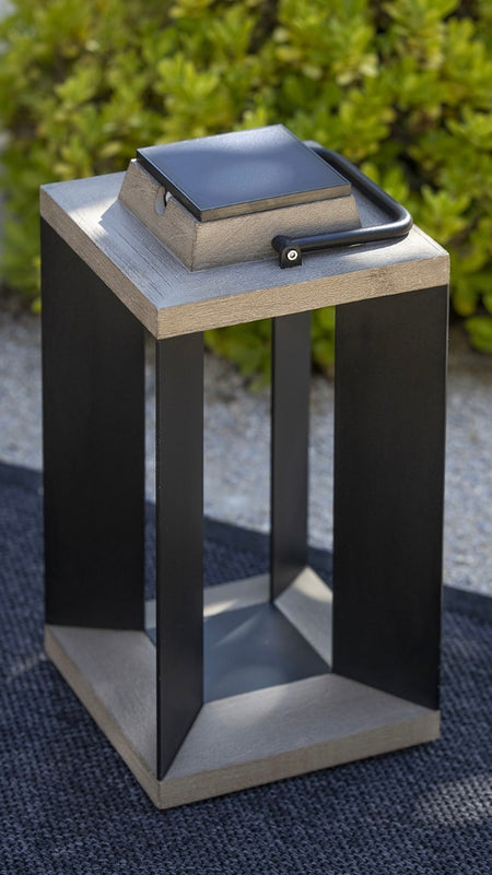 A modern square outdoor solar lantern with black metal frame and teak wood top and base with square solar light on top, set on a dark textured rug with green foliage in the background.