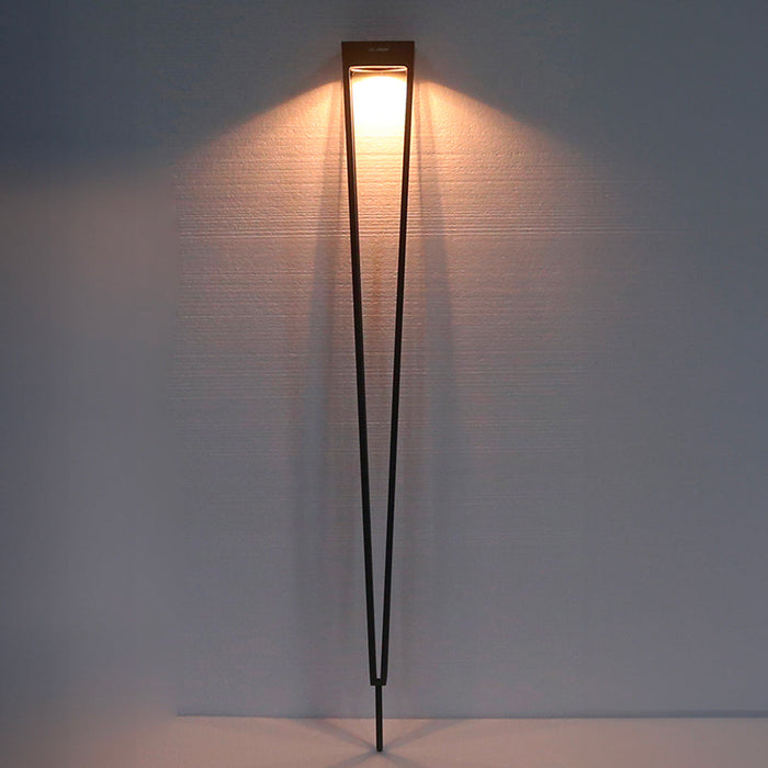 A modern minimalist solar torch with a long, thin black corten aluminium frame and a rectangular warm white light fixture at the top, casting a soft warm glow on a textured gray wall.