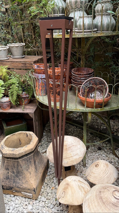 A tall, dark brown metal freestanding lamp or light fixture stands in the center of an outdoor garden or patio display with shelves of ceramic vases, bowls, planters, and wooden decor pieces on a gravel surface.