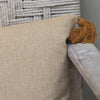 A close-up of a beige and white herringbone patterned throw pillow with a mustard brown tassel, resting on a rustic wooden armchair in front of a woven rope chair back.