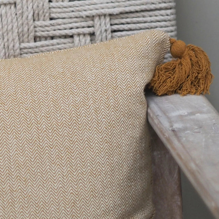 A close-up of a beige and white herringbone patterned throw pillow with a mustard brown tassel, resting on a rustic wooden armchair in front of a woven rope chair back.
